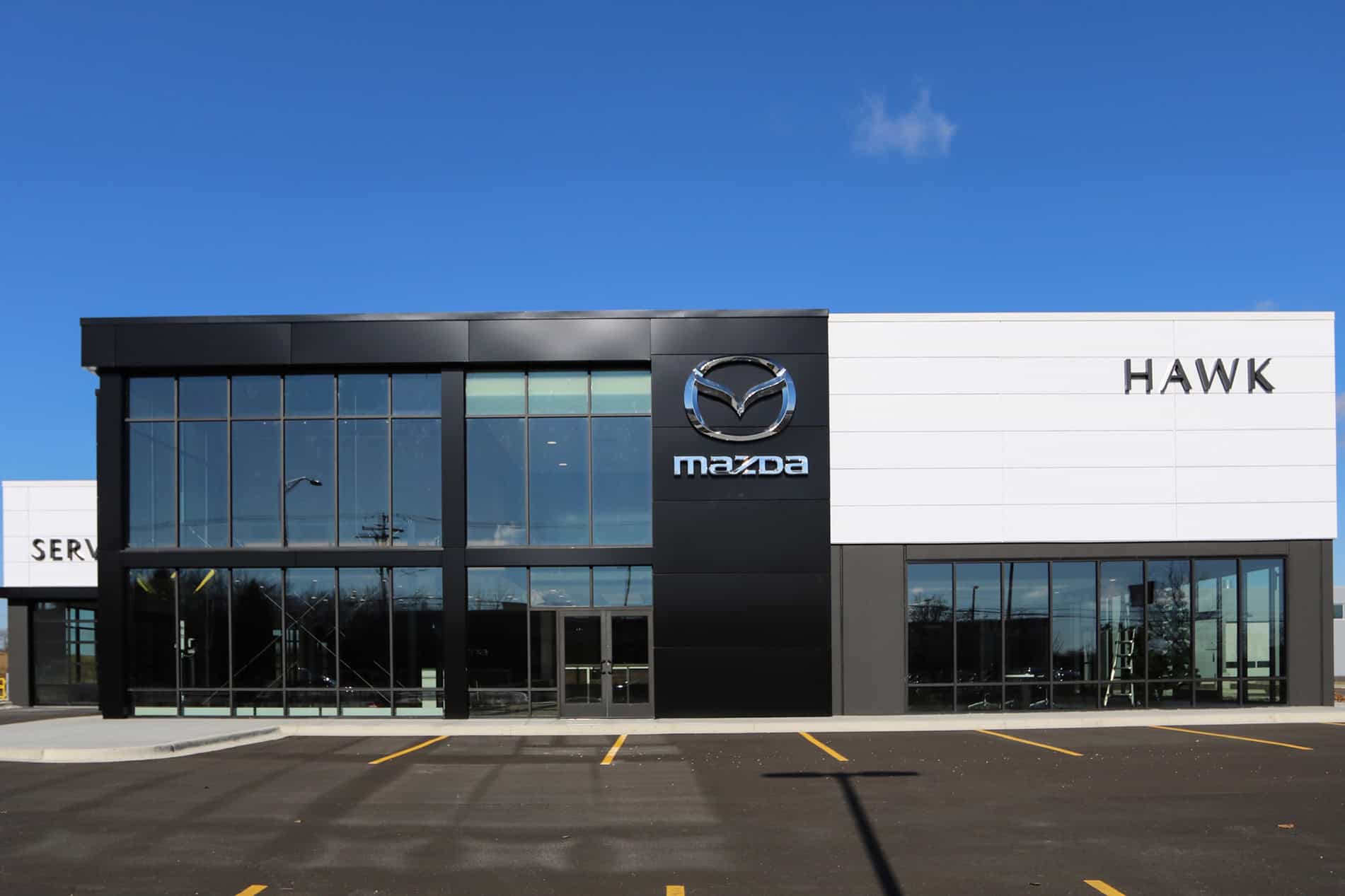 A Mazda sign in the center of the Hawk Auto dealership building, with a two-story glass wall on the left and the name “Hawk” over the office area on the right.