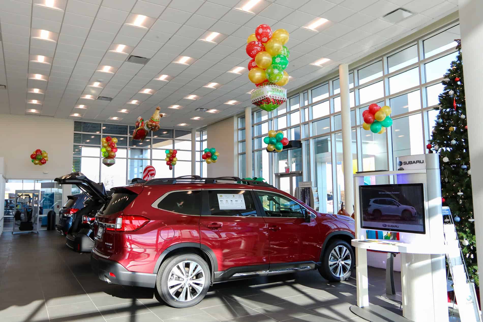 Inside the showroom of the new Hawk Auto dealership, a new red Subaru is parked in a line of other new cars in front of large glass windows adorned with balloons.